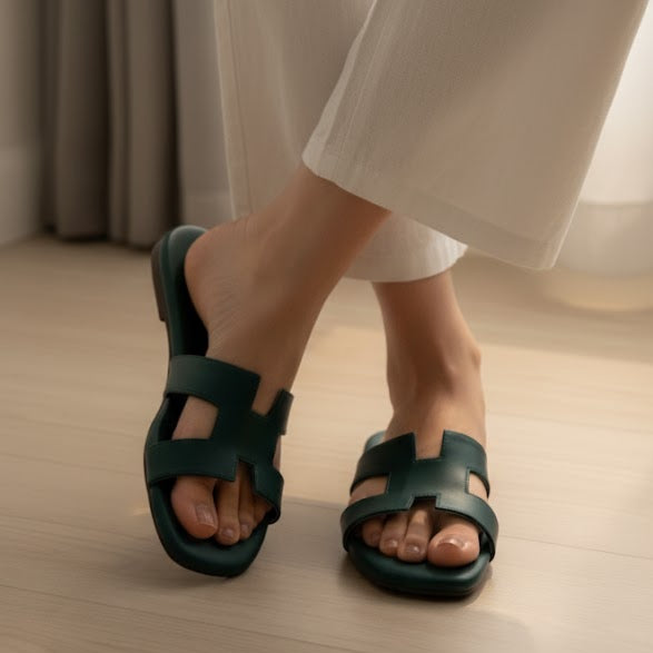 Green sandals worn with white pants on a wooden floor.