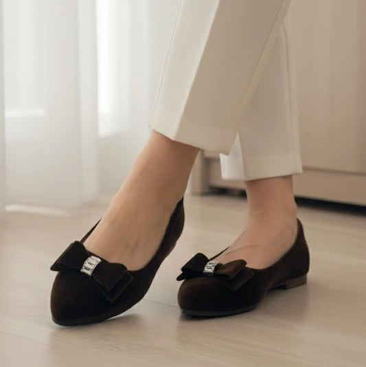 Brown flats with bow details worn with white pants on a light wooden floor.