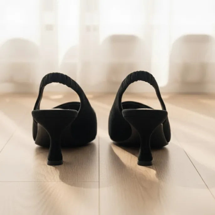 Pair of black high-heeled shoes on a light wooden floor with a blurred curtain background.
