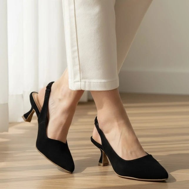 Black high-heeled shoes worn with white pants on a wooden floor.
