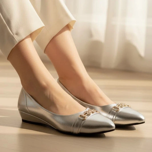 Silver pointed-toe flats worn with white pants on a light wooden floor.