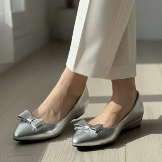 Silver flat shoes with bow details worn by a person on a wooden floor.