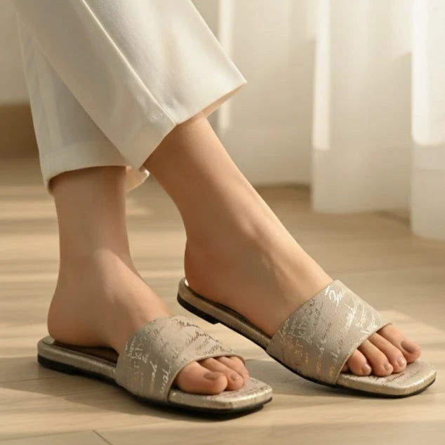 Person wearing light gray slippers on a wooden floor with white pants.