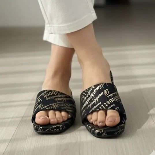 Black slippers with white text worn by a person on a light wooden floor.