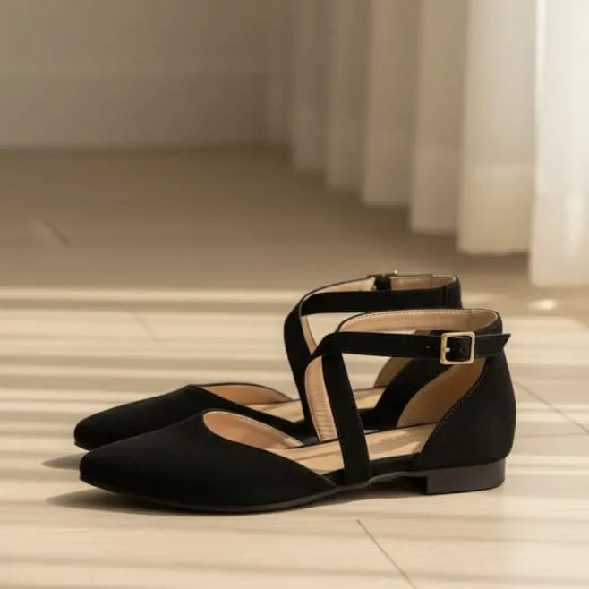 Black flat shoes with thin straps on a light wooden floor.