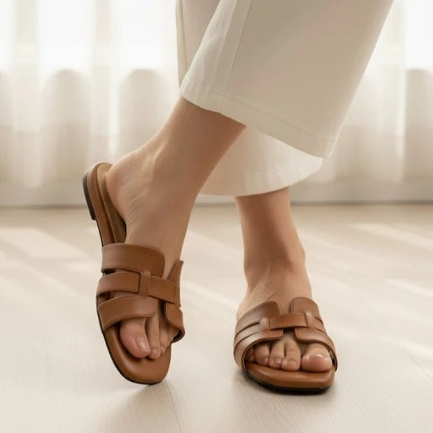 Brown slipper worn with white pants on a light wooden floor.