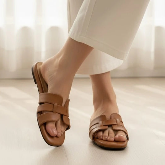 Brown slipper worn with white pants on a light wooden floor.