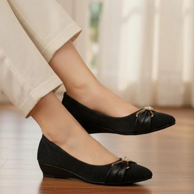 Black flat shoes with decorative bows worn by a person sitting on a wooden floor.