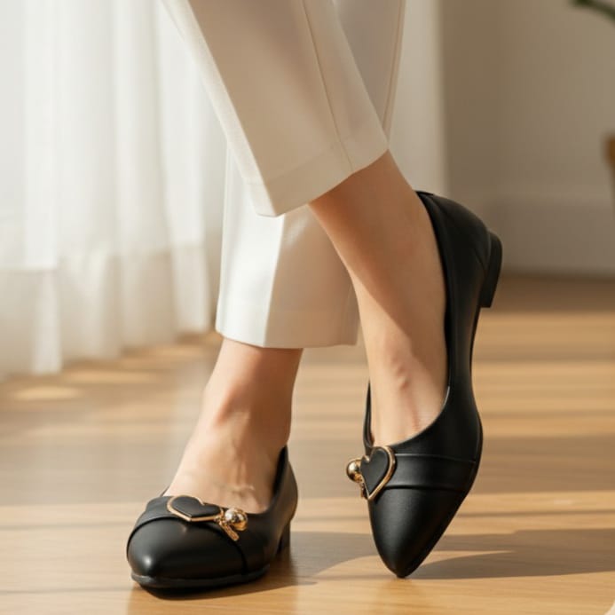Black shoes with heart-shaped gold buckle worn by a person on a wooden floor.