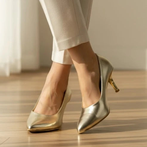 Gold high-heeled shoes worn with light-colored pants on a wooden floor.